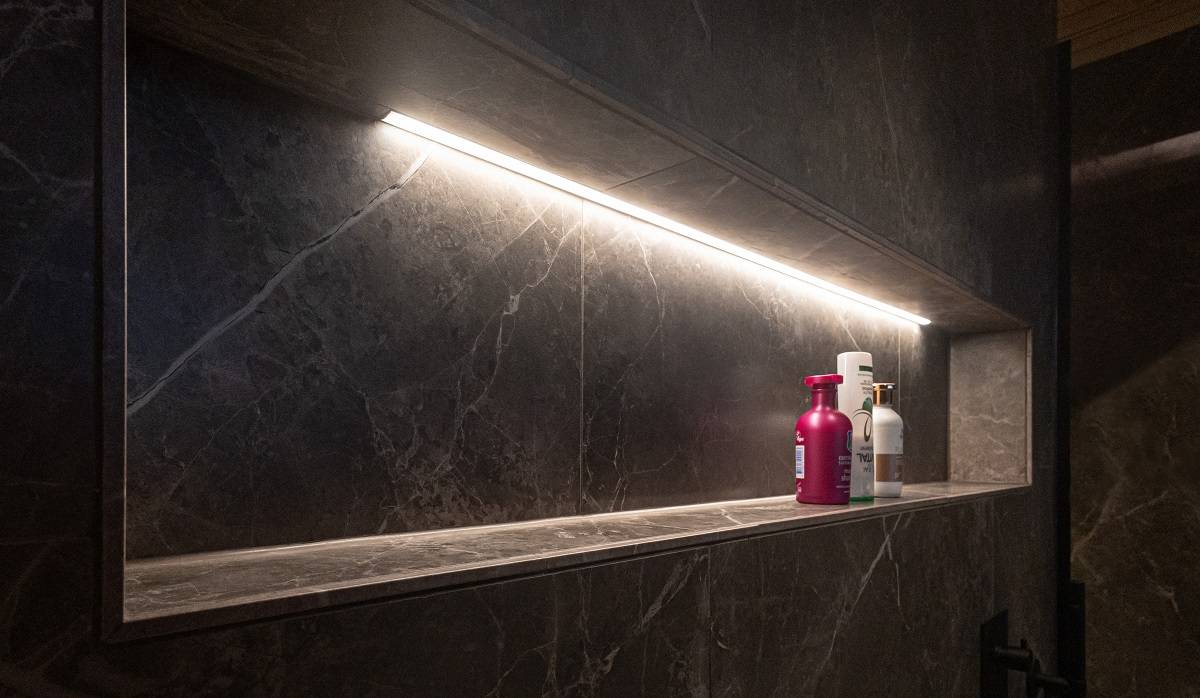 En la pared de piedra gris oscuro de un cuarto de baño, el estante del champú está iluminado por una tira de LED en un perfil de aluminio con una temperatura de color de luz neutra.